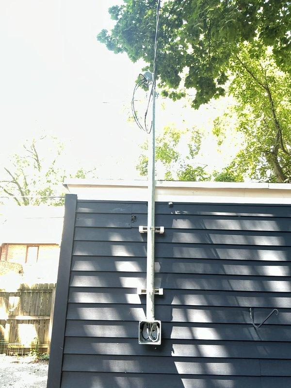 Disconnected existing single strand wiring to garage and terminated wires in existing junction box on side of house, installing half-inch screw-in filler for proper closure. Ran new dedicated circuit from main electrical panel with PVC conduit up exterior wall of residence. Installed point of attachment on house and aerial run to garage using 2-inch rigid conduit with unistrut mounting system to achieve proper clearance from siding and maintain minimum 10-foot height clearance. Mounted screw-on wire holder and transitioned to PVC junction box with Myers hub, converting to Romex in flex conduit to re-feed existing garage junction box. Aerial installation method selected due to large trees in backyard area. Installation includes conduit runs on both structures and overhead wire span between buildings.