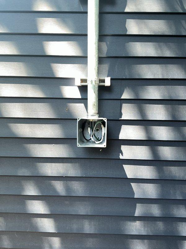Disconnected existing single strand wiring to garage and terminated wires in existing junction box on side of house, installing half-inch screw-in filler for proper closure. Ran new dedicated circuit from main electrical panel with PVC conduit up exterior wall of residence. Installed point of attachment on house and aerial run to garage using 2-inch rigid conduit with unistrut mounting system to achieve proper clearance from siding and maintain minimum 10-foot height clearance. Mounted screw-on wire holder and transitioned to PVC junction box with Myers hub, converting to Romex in flex conduit to re-feed existing garage junction box. Aerial installation method selected due to large trees in backyard area. Installation includes conduit runs on both structures and overhead wire span between buildings.