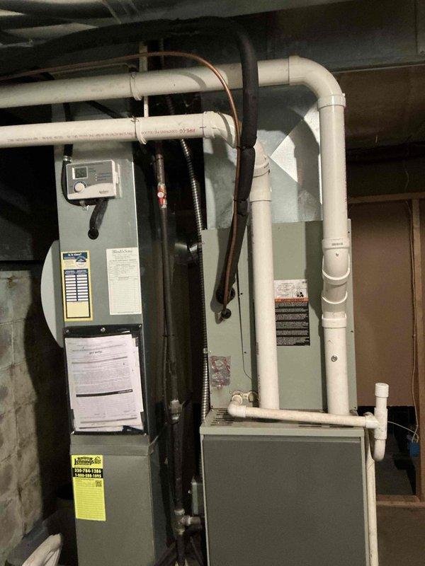 Completed comprehensive HVAC system estimate for furnace and air conditioning replacement in residential basement installation. Evaluated existing gray furnace unit with associated components including white PVC condensate drainage piping, copper refrigerant lines, black electrical conduit, and digital control panel. Documented current system configuration with vertically-mounted thermostat, multiple pipe penetrations through ceiling, and standard basement mechanical room setup with concrete block walls and floor. Photographed all equipment labels, warning stickers, product information sheets, and service documentation for accurate replacement specifications and proper sizing calculations.