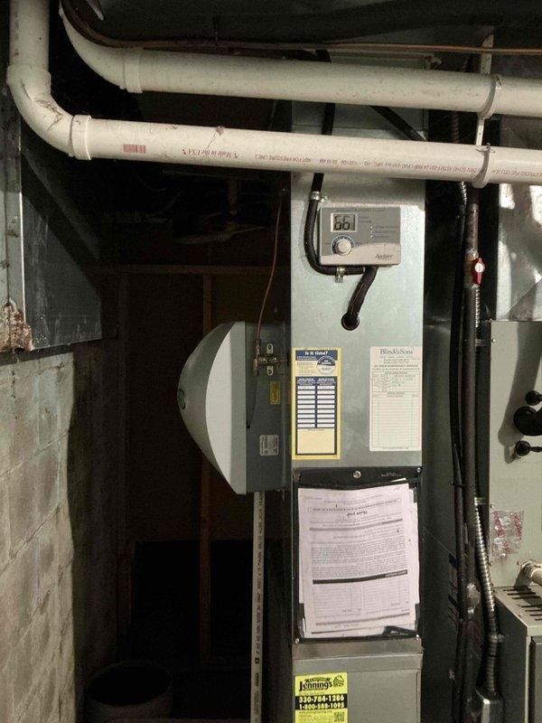 Completed comprehensive HVAC system estimate for furnace and air conditioning replacement in residential basement installation. Evaluated existing gray furnace unit with associated components including white PVC condensate drainage piping, copper refrigerant lines, black electrical conduit, and digital control panel. Documented current system configuration with vertically-mounted thermostat, multiple pipe penetrations through ceiling, and standard basement mechanical room setup with concrete block walls and floor. Photographed all equipment labels, warning stickers, product information sheets, and service documentation for accurate replacement specifications and proper sizing calculations.