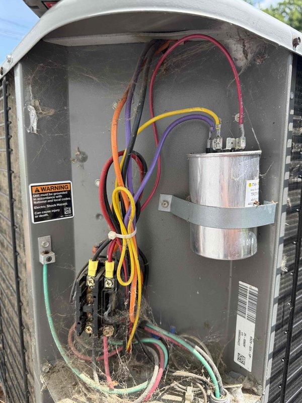 Completed routine MVP cooling maintenance visit on 20-year-old outdoor air conditioning system. Performed comprehensive inspection of cylindrical condenser unit, examining horizontal metal fins for damage and verifying proper airflow through radial fan grille assembly. Inspected electrical panel and control box, checking all wire connections including red, orange, yellow, purple, black, and green conductors secured with proper bundling. Verified capacitor mounting and electrical integrity, noting weathered but functional condition consistent with system age. Examined terminal blocks and connection points for secure attachment and proper operation. Unit operating within normal parameters for age and condition, with condenser positioned appropriately on concrete pad with adequate clearance from structure.