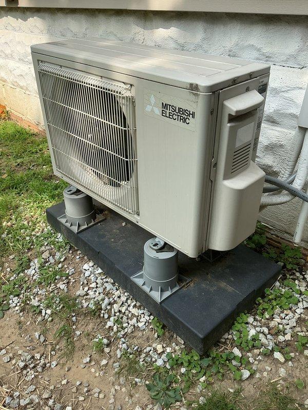 Responded to service call regarding unstable outdoor condenser unit installation. Upon arrival, found Mitsubishi Electric air conditioning condenser improperly leveled on composite pad with inadequate support. Unit was leaning significantly due to settling of ground surface and insufficient base preparation beneath equipment pad. Inspected installation site and identified issues with pad placement on uneven substrate consisting of mixed soil, gravel, and vegetation. Repositioned and leveled condenser unit on existing composite pad, ensuring proper stability and secure mounting. Verified refrigerant lines and electrical conduit connections remained intact and undamaged. Confirmed unit operation and structural stability upon completion.