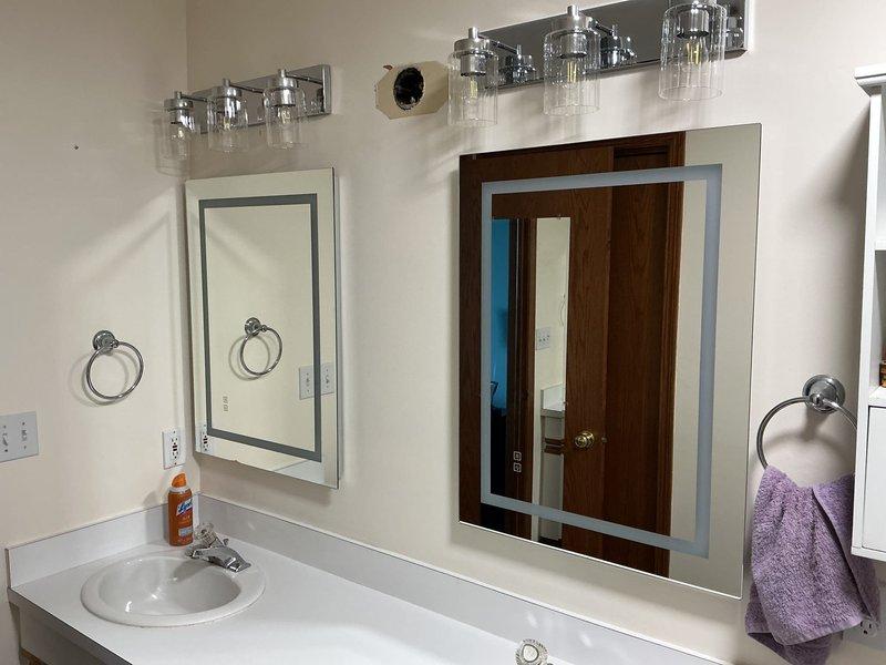 Completed comprehensive electrical upgrades in multiple rooms including bathroom exhaust fan replacements, vanity lighting installations, and ceiling fan work. In the front bathroom, replaced exhaust fan, removed existing vanity light, and installed new vanity light with single gang old work box behind lighted mirror including switched clock outlet for mirror power supply. In the master bathroom, replaced exhaust fan, removed old vanity light, and installed two new vanity lights centered above sinks with two lighted mirrors, utilizing existing switch leg wiring where possible with new runs through attic as needed, and installed switched clock outlets behind each mirror. In the living room, installed light kit to existing ceiling fan and recessed media box behind television for clean wire management. In the master bedroom, replaced ceiling fan with new unit and installed ceiling fan-rated electrical box as required. All customer-provided fixtures including lights, mirrors, exhaust fans,