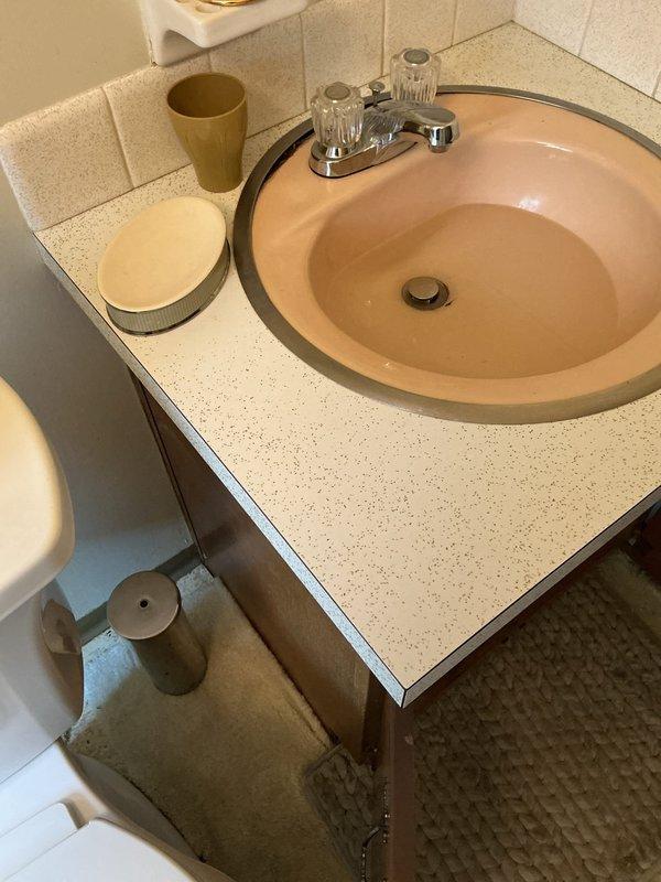 Responded to service call for clogged bathroom sink drain. Upon arrival, assessed the peach-colored vanity sink with chrome dual-handle faucet and inspected the drainage system. Cleared the blockage in the drain line, restoring proper water flow through the basin. Inspection of the cabinet beneath the sink revealed significant pre-existing water damage to the particleboard flooring and back wall, with extensive brown staining and warping indicating long-term moisture exposure. The P-trap and drain assembly connections were examined and found to be properly sealed with no active leaks present at time of service. Drain now functioning properly with water flowing freely through the system.