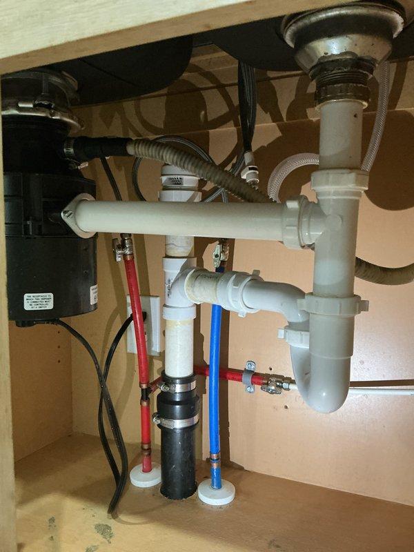Responded to water leak under kitchen sink in 22-year-old residential property. Upon inspection, discovered active leak in PVC drainage assembly beneath sink, with visible water damage and dark staining present on cabinet floor in multiple areas. Plumbing configuration included white PVC P-trap system, garbage disposal unit with connecting drain lines, dishwasher drain connection, and standard hot/cold water supply lines with braided flex hoses. Customer indicated that kitchen pipes had been previously replaced approximately 14 months prior and inquired about potential warranty coverage or service discount. Documented existing conditions with photographs showing full plumbing assembly, drainage connections, water supply lines, and extent of water damage to cabinet interior. Service appointment was rescheduled to allow for warranty verification and proper repair planning.