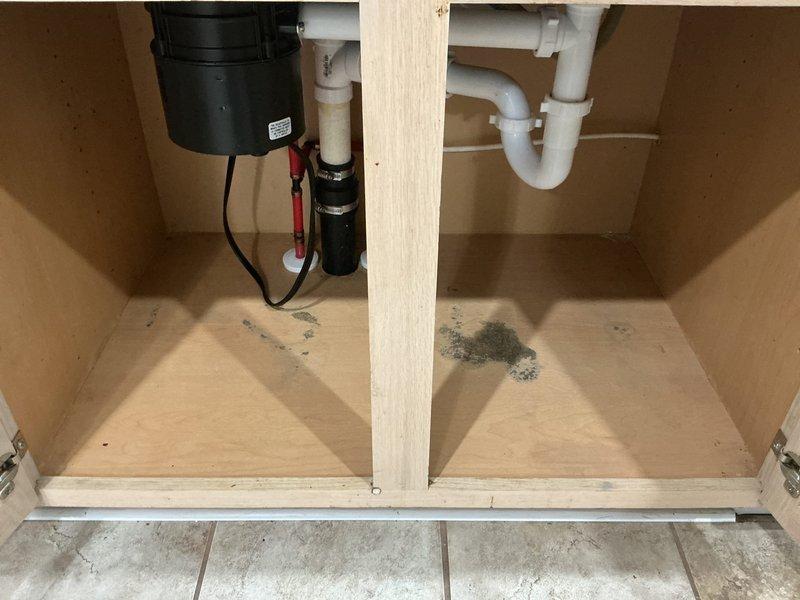 Responded to water leak under kitchen sink in 22-year-old residential property. Upon inspection, discovered active leak in PVC drainage assembly beneath sink, with visible water damage and dark staining present on cabinet floor in multiple areas. Plumbing configuration included white PVC P-trap system, garbage disposal unit with connecting drain lines, dishwasher drain connection, and standard hot/cold water supply lines with braided flex hoses. Customer indicated that kitchen pipes had been previously replaced approximately 14 months prior and inquired about potential warranty coverage or service discount. Documented existing conditions with photographs showing full plumbing assembly, drainage connections, water supply lines, and extent of water damage to cabinet interior. Service appointment was rescheduled to allow for warranty verification and proper repair planning.