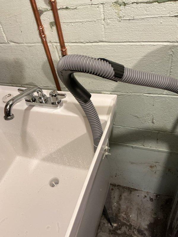 Removed existing laundry tub and installed new single bowl utility sink in basement with deck mount chrome faucet featuring dual handles. Cut existing water lines before original tees and ran new copper supply lines with tees to serve both the faucet and washing machine hookup. Installed new shut-off valves, boiler drains, and washer hoses. Eliminated galvanized standpipe for washing machine and redirected discharge into laundry tub. Connected laundry tub drain to existing drain line. All copper piping properly soldered and secured to wall, with visible vertical runs along painted cinder block wall. Installation completed in utility room setting with concrete floor.