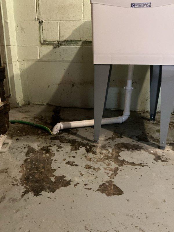 Removed existing laundry tub and installed new single bowl utility sink in basement with deck mount chrome faucet featuring dual handles. Cut existing water lines before original tees and ran new copper supply lines with tees to serve both the faucet and washing machine hookup. Installed new shut-off valves, boiler drains, and washer hoses. Eliminated galvanized standpipe for washing machine and redirected discharge into laundry tub. Connected laundry tub drain to existing drain line. All copper piping properly soldered and secured to wall, with visible vertical runs along painted cinder block wall. Installation completed in utility room setting with concrete floor.
