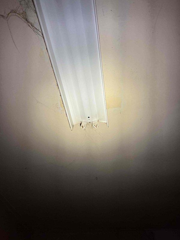 Responded to service call for broken fluorescent socket in laundry room of 40+ year old home where socket failure occurred during bulb replacement attempt. Upon arrival, inspected fluorescent fixture mounted in corner near ceiling, documented existing condition showing signs of age and wear on surrounding surfaces. Verified membership status and applicable service plan coverage prior to proceeding with work. Evaluated socket integrity and determined whether socket replacement alone would suffice or if complete fixture replacement was necessary due to age-related deterioration of internal components. Photographed fixture in both illuminated and detailed views to document pre-existing conditions and justify repair recommendations. Completed necessary repairs to restore proper lighting function in laundry room area.