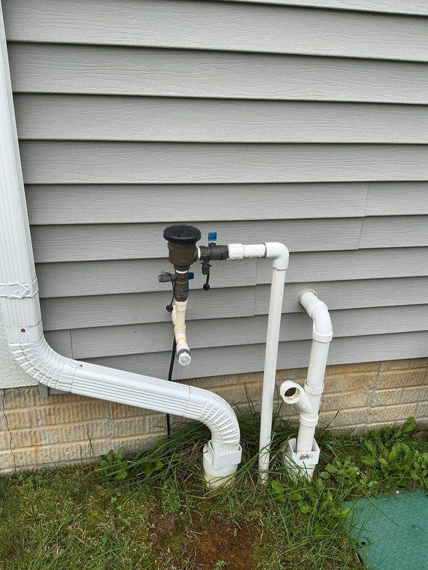 Completed annual backflow preventer testing on exterior residential vacuum breaker assembly located on right side of home. Tested device per industry standards to ensure proper operation and compliance with cross-connection control regulations. Unit passed inspection with all components functioning correctly. Documented installation condition with photographs showing vacuum breaker assembly with blue-handled valve connected to white PVC piping system mounted against building exterior at ground level.