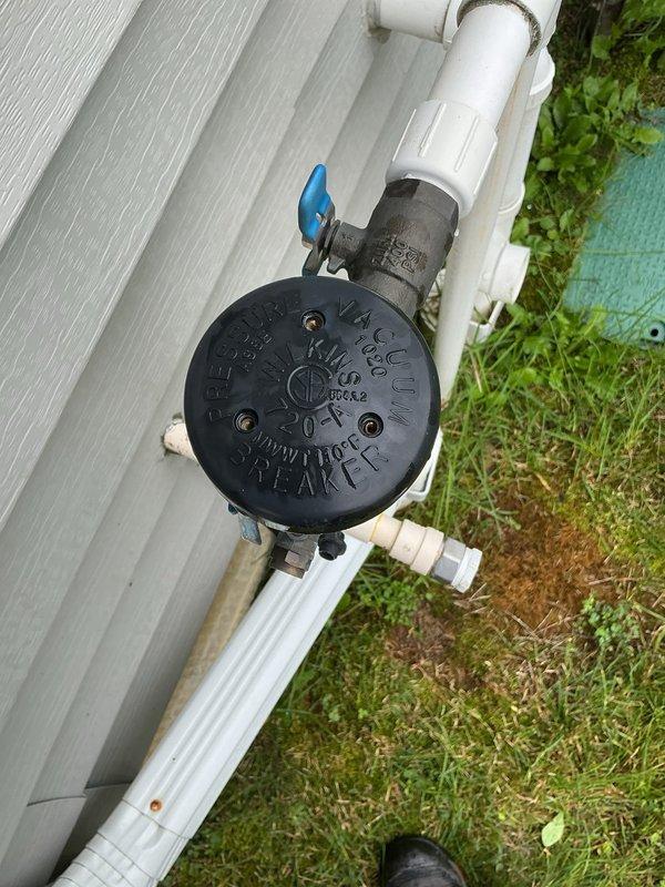 Completed annual backflow preventer testing on exterior residential vacuum breaker assembly located on right side of home. Tested device per industry standards to ensure proper operation and compliance with cross-connection control regulations. Unit passed inspection with all components functioning correctly. Documented installation condition with photographs showing vacuum breaker assembly with blue-handled valve connected to white PVC piping system mounted against building exterior at ground level.