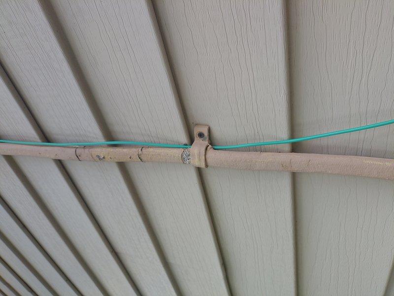Inspected and documented AC circuit routing along exterior building structure. Identified turquoise-colored electrical wire running through mounting bracket system on wooden beam with visible age and wear. Verified circuit pathway where wire crosses insulated pipe section showing deteriorated foam insulation with exposed underlayment. Photographed installation for service records showing wire management and mounting hardware configuration on textured paneling surface.