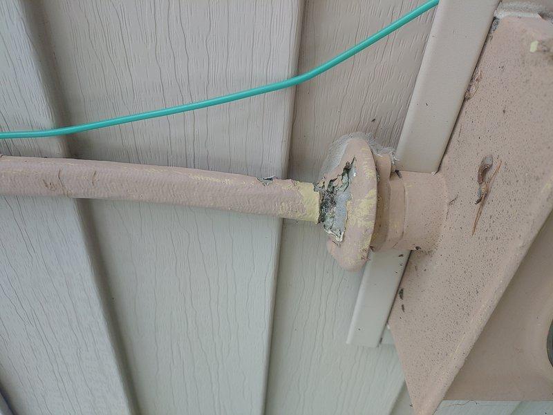 Inspected and documented AC circuit routing along exterior building structure. Identified turquoise-colored electrical wire running through mounting bracket system on wooden beam with visible age and wear. Verified circuit pathway where wire crosses insulated pipe section showing deteriorated foam insulation with exposed underlayment. Photographed installation for service records showing wire management and mounting hardware configuration on textured paneling surface.