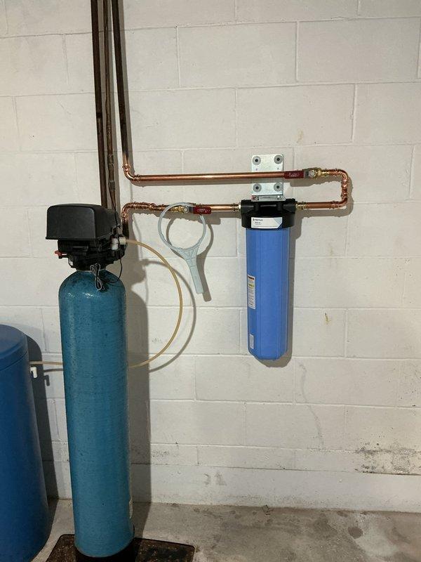Installed whole home water filtration system to address sediment issues in the residential water supply. The installation included mounting a blue sediment filter housing with cartridge on the existing copper plumbing line, integrated with the current water treatment equipment including the existing softener tank. All components were properly secured to the concrete block wall with appropriate mounting brackets, connections were made using copper piping, and the system was tested for proper operation and flow.