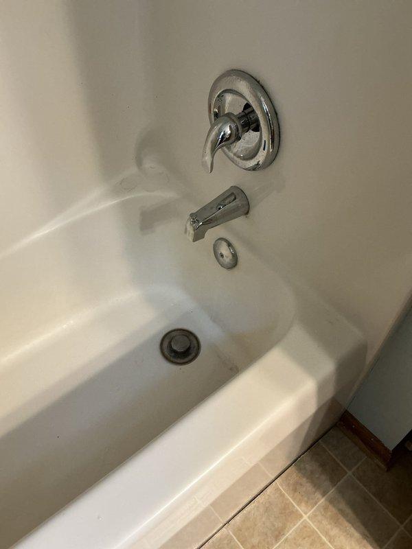 Responded to water leak complaint originating from upstairs bathroom and penetrating through ceiling below. Gained access to plumbing chase through existing access panel to inspect drain assembly and supply lines. Upon investigation, discovered corroded copper supply lines with visible mineral deposits and oxidation, indicating long-term moisture exposure. Identified compromised drain connections at bathtub fixture as primary source of leak. Documented existing conditions including deteriorated pipe surfaces, accessible shut-off valves with red handles, and white PVC drain line with signs of age-related wear. Photographed bathtub fixture assembly showing chrome-finished faucet, tub spout with diverter, and drain components for reference. Inspected wall cavity revealing copper supply pipes and drain plumbing requiring repair or replacement to resolve active leak condition.