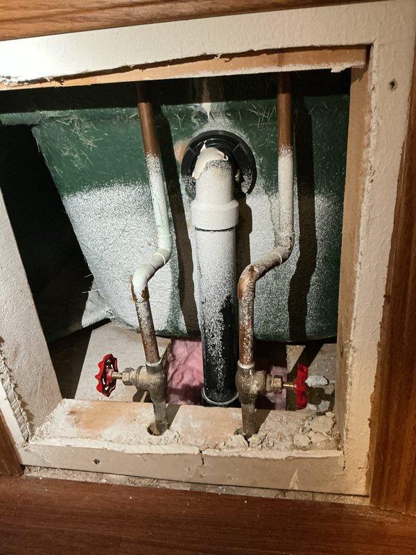 Responded to water leak complaint originating from upstairs bathroom and penetrating through ceiling below. Gained access to plumbing chase through existing access panel to inspect drain assembly and supply lines. Upon investigation, discovered corroded copper supply lines with visible mineral deposits and oxidation, indicating long-term moisture exposure. Identified compromised drain connections at bathtub fixture as primary source of leak. Documented existing conditions including deteriorated pipe surfaces, accessible shut-off valves with red handles, and white PVC drain line with signs of age-related wear. Photographed bathtub fixture assembly showing chrome-finished faucet, tub spout with diverter, and drain components for reference. Inspected wall cavity revealing copper supply pipes and drain plumbing requiring repair or replacement to resolve active leak condition.