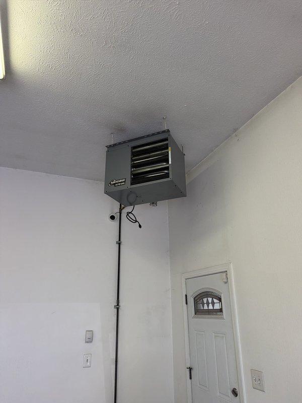 Installed customer-supplied garage heater unit in residential garage space. Mounted gray metal heating unit to upper wall corner near ceiling with secure mounting brackets, ran black electrical conduit vertically down wall for power supply, and completed all electrical connections to ensure proper operation of the wall-mounted heating system.