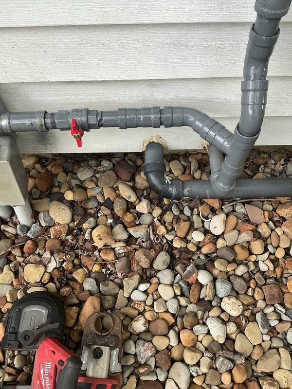 Installed exterior gas line test setup with gray PVC piping configuration including horizontal and vertical runs, incorporated red-handled ball valve for system control, and secured all components with proper pipe straps and supports against building exterior. Installation completed over decorative river rock ground cover with all connections, T-joints, and elbow fittings properly assembled and tested for system integrity.