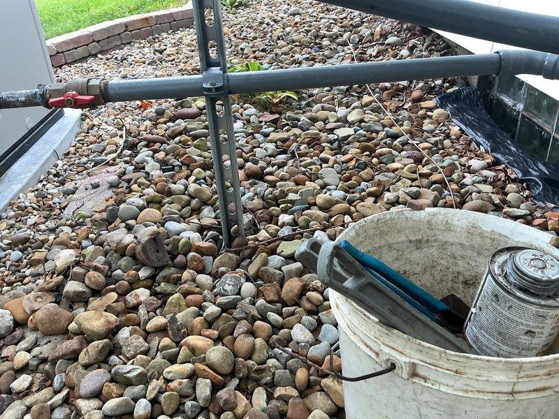 Installed exterior gas line test setup with gray PVC piping configuration including horizontal and vertical runs, incorporated red-handled ball valve for system control, and secured all components with proper pipe straps and supports against building exterior. Installation completed over decorative river rock ground cover with all connections, T-joints, and elbow fittings properly assembled and tested for system integrity.