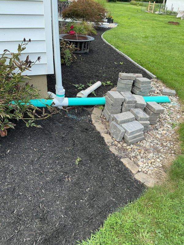 Replaced existing sewage pit and sewage pump with new units, connecting new pump to existing discharge line and vent system. Modified lavatory drain configuration for basement bathroom by connecting to sewage pit vent rather than direct pit lid connection, requiring access through drywall and tile in basement bathroom area. Jackhammered concrete near existing sewage pit location to install new storm water crock and connected existing perforated pipe to new discharge line running to existing downspout. Installed new 1/3 HP sump pump in new sump crock with new check valve. Tested all connections and discharge systems to verify proper operation and absence of leaks. Repoured concrete to rough finish over excavated areas. Exterior drainage system now includes visible turquoise PVC piping running through foundation planting bed, connecting to white downspout along house siding for proper storm water management.