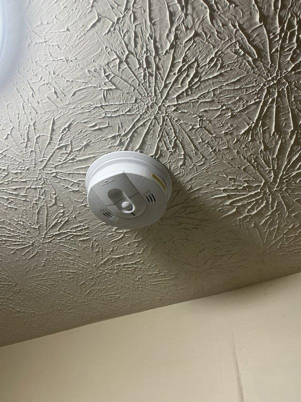 Responded to service call regarding chirping smoke detector near bedroom door. Upon arrival, inspected ceiling-mounted smoke alarm unit and confirmed customer had attempted battery replacement but was unable to properly install new batteries. Assessed smoke detector installation and battery compartment accessibility. Opened detector housing, removed old batteries, and installed fresh batteries with correct polarity. Verified secure battery connection and proper seating within compartment. Tested smoke detector functionality to confirm chirping issue was resolved and unit was operating correctly. Reinstalled detector cover and ensured proper mounting to textured ceiling surface.