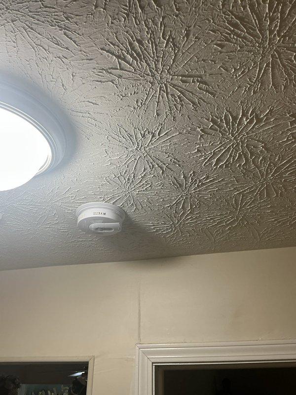 Responded to service call regarding chirping smoke detector near bedroom door. Upon arrival, inspected ceiling-mounted smoke alarm unit and confirmed customer had attempted battery replacement but was unable to properly install new batteries. Assessed smoke detector installation and battery compartment accessibility. Opened detector housing, removed old batteries, and installed fresh batteries with correct polarity. Verified secure battery connection and proper seating within compartment. Tested smoke detector functionality to confirm chirping issue was resolved and unit was operating correctly. Reinstalled detector cover and ensured proper mounting to textured ceiling surface.