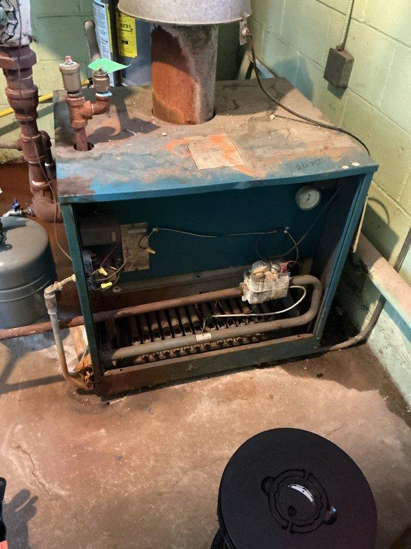 Responded to service call for aging steam gas boiler system with reported water leak from piping. Upon arrival, conducted thorough inspection of the boiler system, which exhibited significant age-related deterioration including extensive rust and corrosion on the expansion tank, worn cabinet surfaces, and oxidized copper piping connections. Located active water leak at pipe connection point and documented system condition through photographic evidence. The boiler cabinet showed faded paint, rust staining on top surfaces, and visible wear throughout, while the cylindrical expansion tank displayed considerable orange-brown rust discoloration on lower portions. Inspected all plumbing connections, valves, fittings, and pressure gauge for additional concerns. Examined burner assembly and electrical wiring visible through open cabinet panel. Assessed overall system integrity and noted multiple components showing advanced stages of deterioration. Provided customer with findings regarding the 