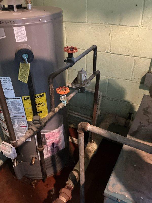 Responded to service call for aging steam gas boiler system with reported water leak from piping. Upon arrival, conducted thorough inspection of the boiler system, which exhibited significant age-related deterioration including extensive rust and corrosion on the expansion tank, worn cabinet surfaces, and oxidized copper piping connections. Located active water leak at pipe connection point and documented system condition through photographic evidence. The boiler cabinet showed faded paint, rust staining on top surfaces, and visible wear throughout, while the cylindrical expansion tank displayed considerable orange-brown rust discoloration on lower portions. Inspected all plumbing connections, valves, fittings, and pressure gauge for additional concerns. Examined burner assembly and electrical wiring visible through open cabinet panel. Assessed overall system integrity and noted multiple components showing advanced stages of deterioration. Provided customer with findings regarding the 