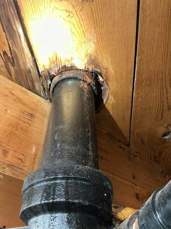 Responded to service call for water leak in basement with unknown source. Upon investigation, traced leak to bathroom above basement area. Inspection revealed compromised toilet flange connection at floor penetration point, with evidence of long-term moisture exposure causing deterioration to surrounding subfloor structure. The wooden subfloor around the toilet drain pipe showed significant water damage and charring effect from prolonged exposure. Documented conditions with photos showing bathroom fixture location and underside floor damage at pipe penetration. Advised customer of findings and recommended complete toilet removal, flange replacement, and subfloor repair to address water infiltration issue and prevent further structural damage to basement ceiling below.