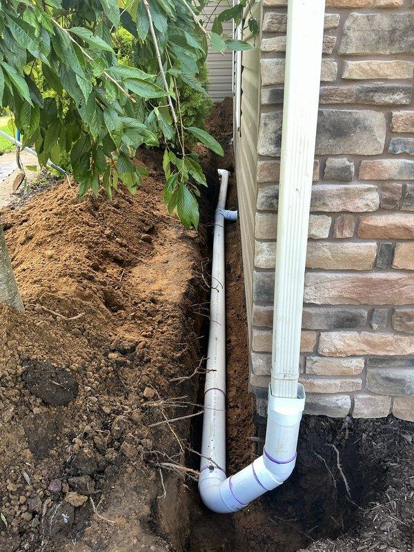 Installed new 4" downspout drainage system for front of residence, connecting garage drains, front door downspout, and corner garage downspout. Trenched new drainage lines from building foundation to front ditch, tunneling under sidewalk where necessary. Daylighted discharge pipe at ditch terminus with gravel surround for erosion control. All white PVC piping installed with proper slope and secure connections to existing downspout system to direct water away from foundation.