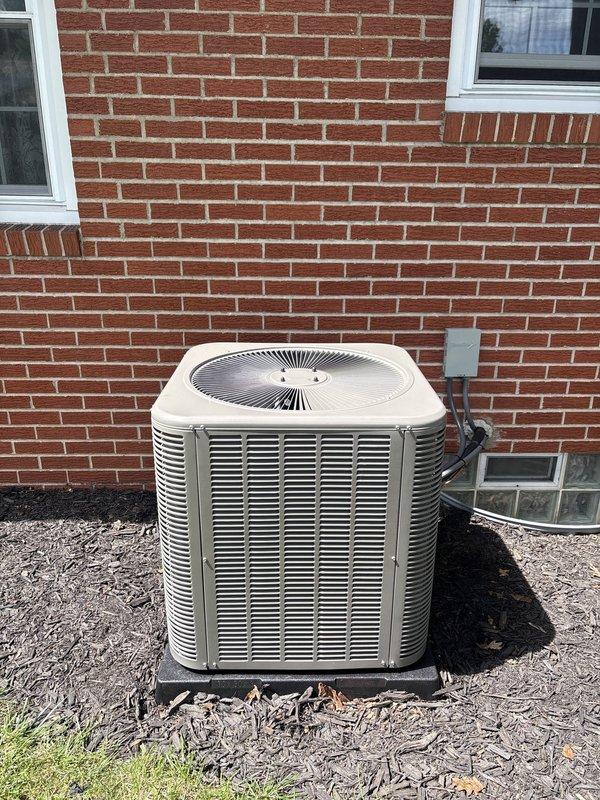 Completed installation of new air conditioning system including outdoor condensing unit and indoor evaporator coil. Outdoor unit was positioned on concrete pad against exterior brick wall with proper clearances maintained, electrical disconnect box mounted to structure, and line set connected between indoor and outdoor components. Indoor evaporator coil was installed in conjunction with existing Trane furnace in basement utility area, with refrigerant lines and condensate drainage properly routed through PVC piping. System was pressure tested for leaks, evacuated to manufacturer specifications, charged with appropriate refrigerant, and cycled through operational testing to verify proper cooling performance and airflow.