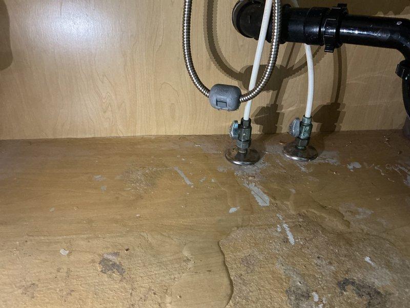 Responded to a water leak under the laundry room sink and discovered significant water damage to the particle board cabinet floor, with visible staining, discoloration, and mineral deposits indicating prolonged moisture exposure. Inspected the plumbing connections, including the disposal assembly, flexible supply lines, and shut-off valves to identify the source of the leak. Documented the extent of the water damage to the cabinet base for repair recommendations.
