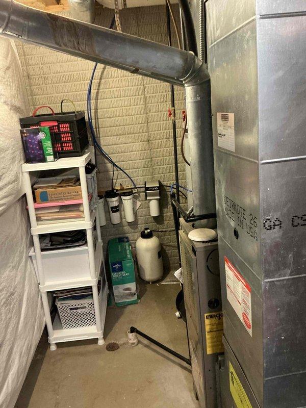 Performed on-site evaluation of 25-year-old residential air conditioning system for replacement estimate. Inspected exterior cylindrical condenser unit with beige housing and vertical louvers, noting age and condition of equipment mounted on concrete pad adjacent to home's exterior. Documented basement furnace and HVAC infrastructure, including ductwork, electrical connections, and existing system components to assess compatibility requirements for new AC installation. Captured photographic documentation of current equipment configuration and installation location for replacement planning purposes.