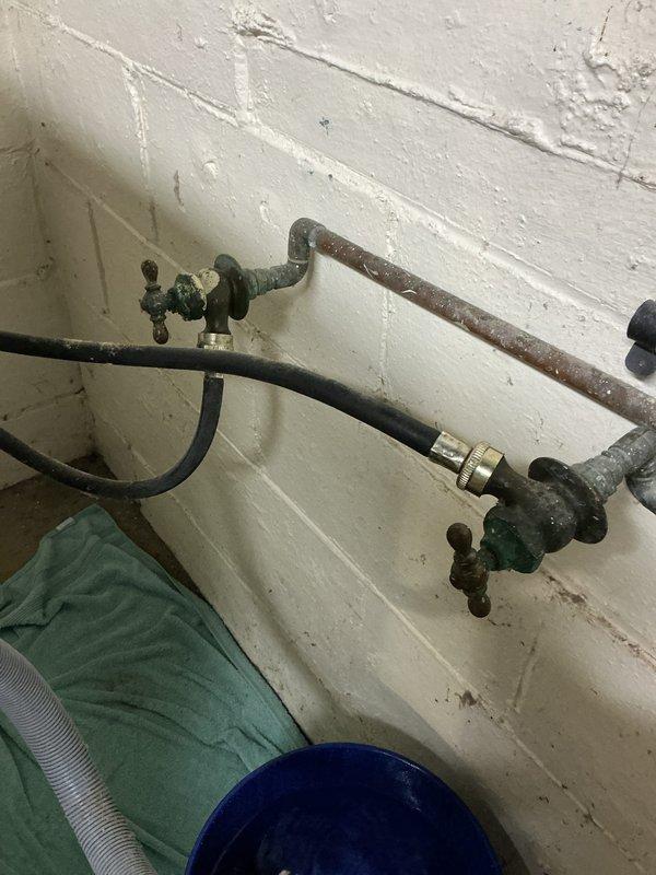Inspected and assessed corroded washer hookup valves and associated plumbing prior to new appliance installation. Found frozen shut-off valve with significant corrosion and green patina on brass components, causing minor drip when operated. Documented extensive deterioration on metal piping including surface rust and oxidation on horizontal supply lines. Identified mixed-age plumbing system with newer PVC and blue plastic piping connected to older corroded metal components. Photographed valve assemblies, pipe conditions, and wall-mounted fixtures showing rust staining and water damage on surrounding surfaces for customer review and repair recommendations.