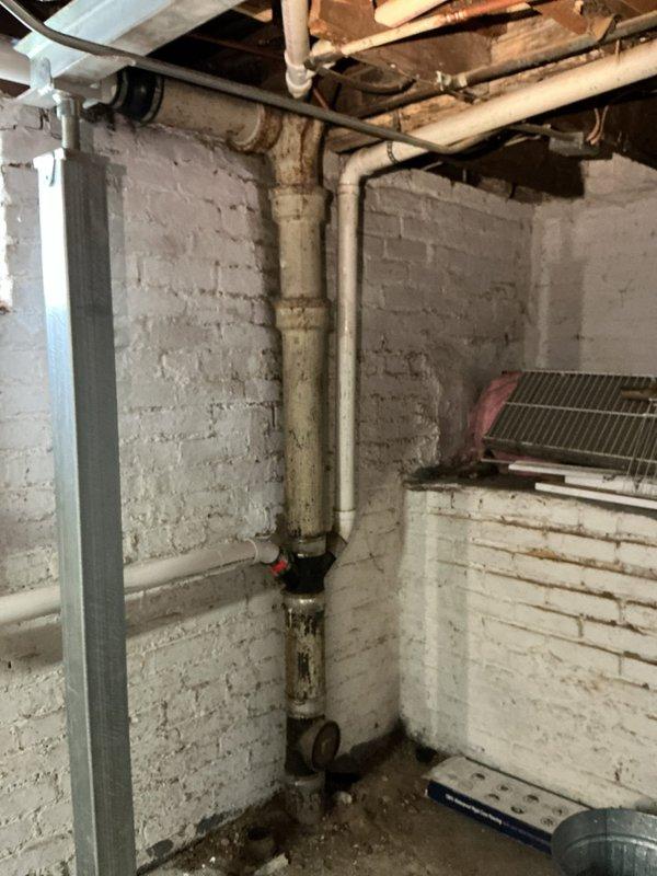 Responded to a main line repair call where a hole was discovered in the drain pipe connecting the toilet to the main plumbing stack. Upon inspection of the basement area, identified the damaged section of pipe along with extensive rust staining and water damage on the foundation walls and floor joists, indicating long-term moisture infiltration from the compromised pipe. The affected area showed significant deterioration with visible staining concentrated around the pipe penetrations and corner sections.

Performed diagnostic evaluation of the drainage system, documenting the condition of both the damaged pipe and the aging cast iron plumbing stack. Photographed the existing plumbing infrastructure, including white PVC drain lines, copper supply lines, and the deteriorated iron stack showing signs of corrosion and age-related wear. The inspection revealed multiple pipe connections and fittings in the basement ceiling area, with evidence of previous water damage and staining on wooden j