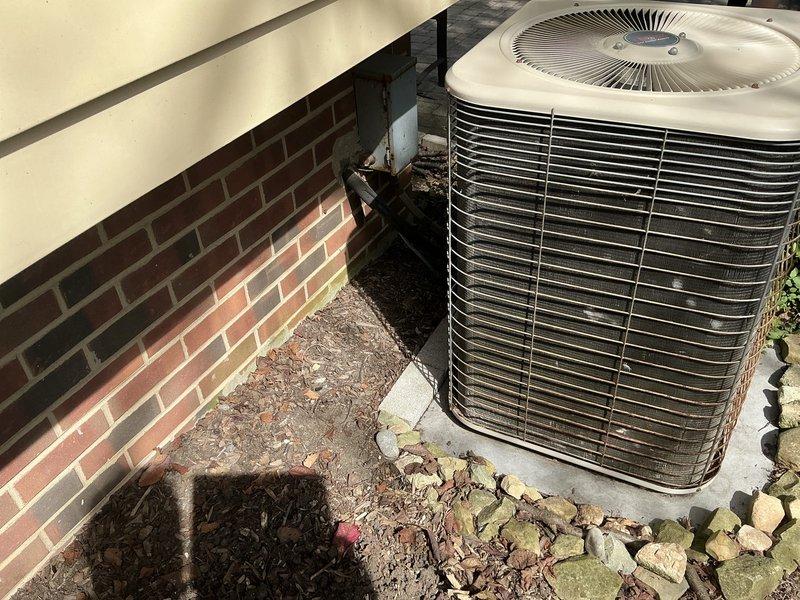 Conducted site evaluation of 30-year-old residential air conditioning system for replacement estimate. Inspected outdoor condensing unit positioned on concrete pad adjacent to brick foundation, noting weathered cylindrical unit with horizontal fins and top-mounted fan grille showing signs of age and outdoor exposure. Documented existing equipment configuration, refrigerant line routing with visible service valve, and installation location constraints between building wall and yard area. Photographed unit condition and surrounding site conditions including mulch ground cover and clearance space. Provided customer with available manufacturer options and system recommendations for complete HVAC replacement.