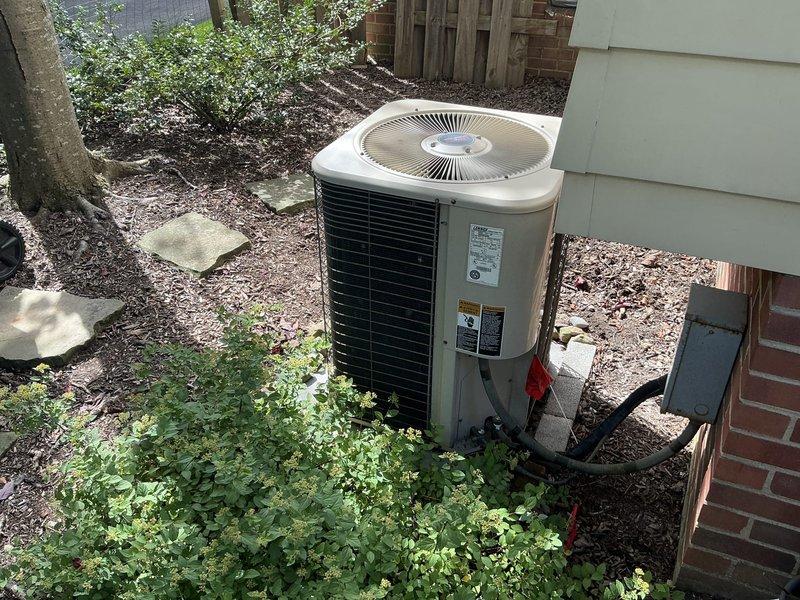 Conducted site evaluation of 30-year-old residential air conditioning system for replacement estimate. Inspected outdoor condensing unit positioned on concrete pad adjacent to brick foundation, noting weathered cylindrical unit with horizontal fins and top-mounted fan grille showing signs of age and outdoor exposure. Documented existing equipment configuration, refrigerant line routing with visible service valve, and installation location constraints between building wall and yard area. Photographed unit condition and surrounding site conditions including mulch ground cover and clearance space. Provided customer with available manufacturer options and system recommendations for complete HVAC replacement.