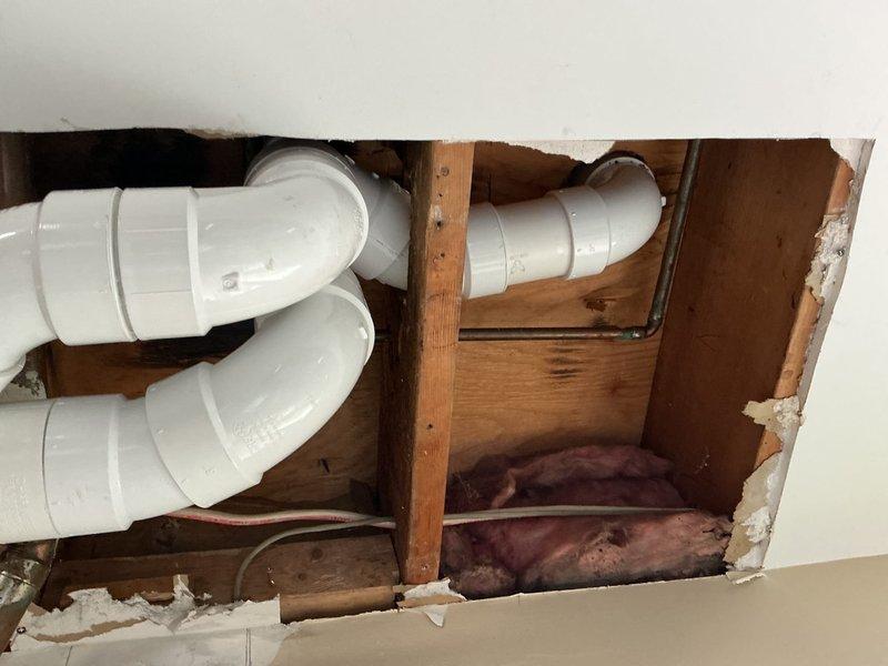 Removed and replaced defective copper double sanitary tee serving back-to-back toilet configuration. Pulled master bathroom toilet and cut open hall bathroom wall to access existing drain and vent stack. Removed horizontal copper piping from kitchen wall connection through both toilet flanges. Installed new 3" PVC drainage system including two new toilet flanges connected to double wye fitting for back-to-back toilet waste lines, then reconnected to existing copper line at kitchen wall. New piping installed at slightly lower elevation than original configuration.