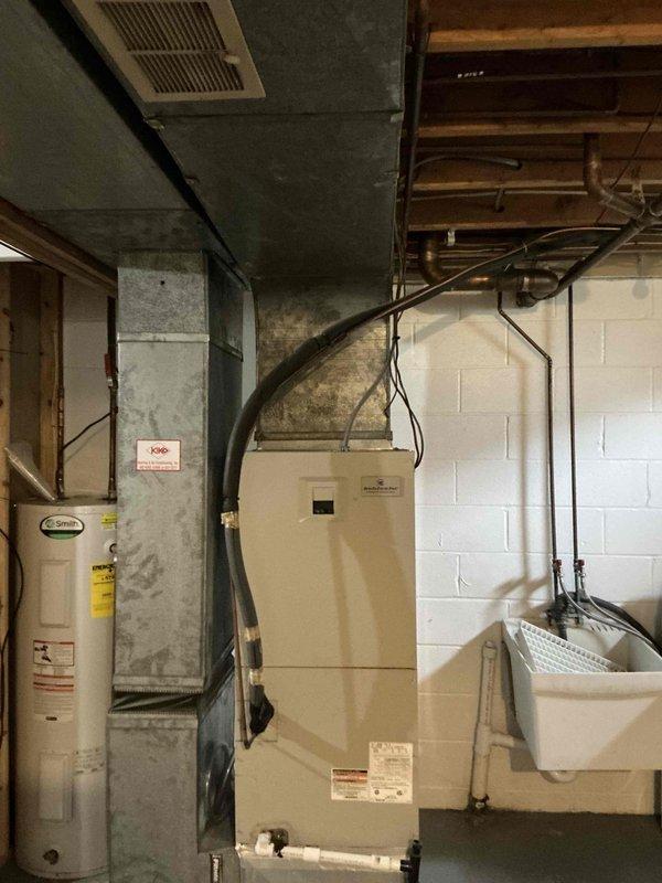 Conducted comprehensive on-site evaluation for complete HVAC system replacement at residential property. Assessed existing cylindrical outdoor condensing unit positioned on concrete pad with platform bases in side yard area, noting current equipment configuration and clearances relative to foundation, privacy fencing, and utility connections. Evaluated indoor air handler and furnace system located in basement mechanical room, documenting current beige cabinet unit alongside water heater, existing ductwork routing through floor joists, and electrical service connections. Photographed both exterior and interior equipment installations to document pre-replacement conditions and facilitate accurate equipment sizing and replacement planning for forthcoming system installation.
