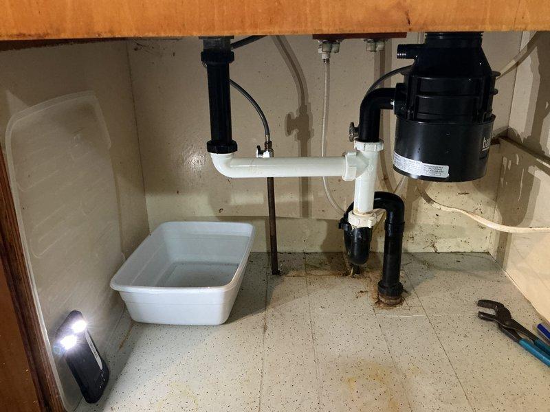 Responded to service call for kitchen sink that would not drain. Upon inspection, found the drainage system under the sink was impeding water flow. Accessed the cabinet beneath the double-basin stainless steel sink and inspected the PVC trap assembly and garbage disposal connections. Cleared the obstruction from the drain line, tested both basins for proper drainage, and verified all plumbing connections were secure and leak-free. System restored to full functionality with water draining properly from both sink basins.