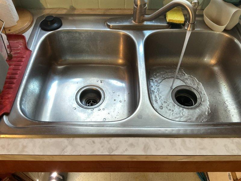 Responded to service call for kitchen sink that would not drain. Upon inspection, found the drainage system under the sink was impeding water flow. Accessed the cabinet beneath the double-basin stainless steel sink and inspected the PVC trap assembly and garbage disposal connections. Cleared the obstruction from the drain line, tested both basins for proper drainage, and verified all plumbing connections were secure and leak-free. System restored to full functionality with water draining properly from both sink basins.