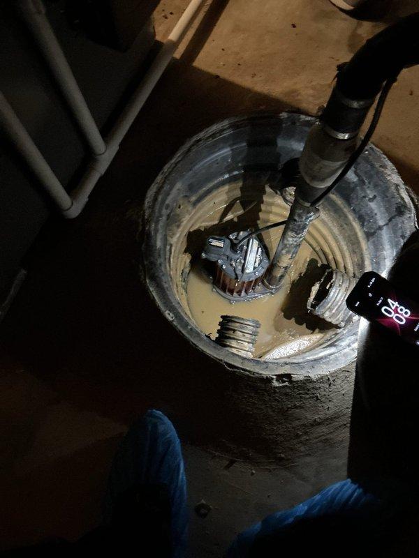 Responded to service call for non-operational sump pump system in basement. Upon arrival, inspected submersible sump pump installed in corrugated plastic basin and found unit experiencing operational failure. Pump showed visible wear with sediment and mineral deposit accumulation around housing base, indicating extended service life. Evaluated discharge piping, electrical connections, and float mechanism. Documented existing moisture concerns in basement area, including water staining on concrete floor and efflorescence on block walls indicating historical water infiltration issues. Noted electrical wiring configuration and plumbing system connections for sump discharge. Photographed current system condition including pump placement, basin configuration, and surrounding utility area for service records.