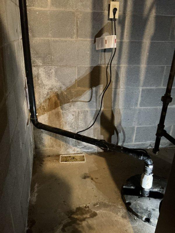 Responded to service call for non-operational sump pump system in basement. Upon arrival, inspected submersible sump pump installed in corrugated plastic basin and found unit experiencing operational failure. Pump showed visible wear with sediment and mineral deposit accumulation around housing base, indicating extended service life. Evaluated discharge piping, electrical connections, and float mechanism. Documented existing moisture concerns in basement area, including water staining on concrete floor and efflorescence on block walls indicating historical water infiltration issues. Noted electrical wiring configuration and plumbing system connections for sump discharge. Photographed current system condition including pump placement, basin configuration, and surrounding utility area for service records.