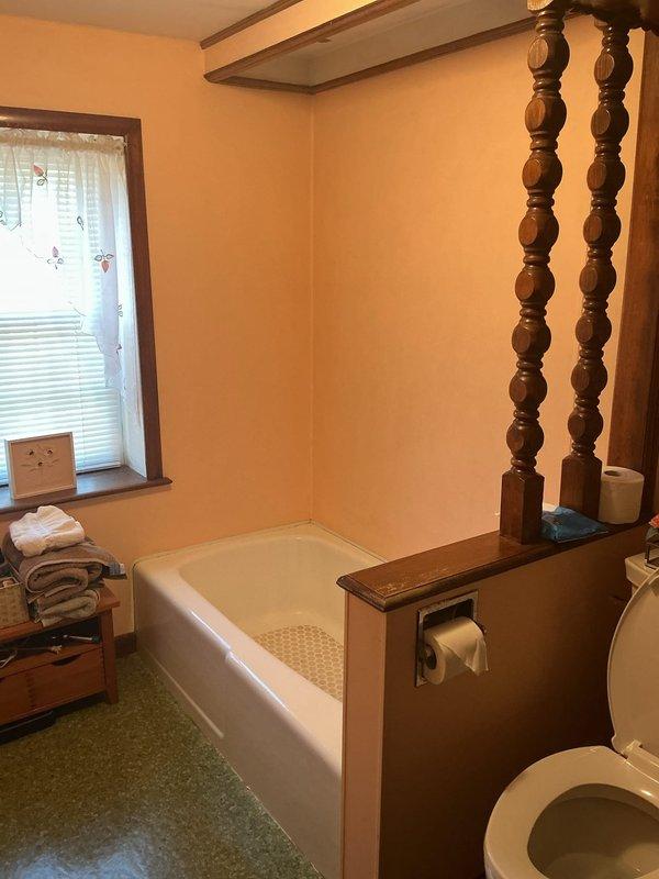Converted existing bathtub to walk-in shower installation. Removed standard tub fixture, decorative wooden posts, and associated hardware from corner bathroom location. Prepared plumbing connections and drainage system for new shower configuration. Documented existing bathroom conditions including yellow-painted walls, wooden vanity, and speckled flooring prior to conversion work.