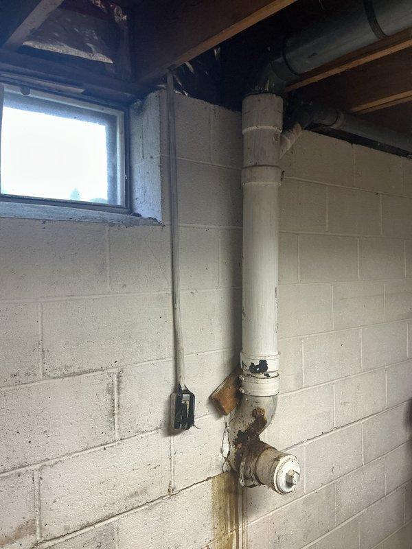 Responded to water leak in basement beneath upstairs bathroom with approximately 6-foot diameter pooling area. Investigated overhead plumbing system to identify drip source from recently remodeled bathroom installation completed one week prior. Inspected white PVC drainage assembly mounted on concrete block wall, discovering significant rust staining and corrosion around elbow joint and valve connection points, with visible deterioration and water damage patterns indicating active leak at fitting. Documented extensive rust discoloration, mineral deposits, and structural concerns including wall cracks near wooden support block mounting. Additionally assessed 4-inch cast iron main drain line penetrating concrete block foundation to septic system, noting advanced scaling and rust deterioration requiring replacement consideration. Photographed all affected plumbing components, leak evidence, and deteriorating infrastructure for repair recommendations. Advised that both the active overhead 