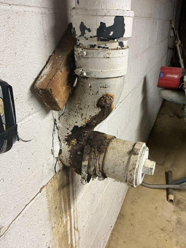 Responded to water leak in basement beneath upstairs bathroom with approximately 6-foot diameter pooling area. Investigated overhead plumbing system to identify drip source from recently remodeled bathroom installation completed one week prior. Inspected white PVC drainage assembly mounted on concrete block wall, discovering significant rust staining and corrosion around elbow joint and valve connection points, with visible deterioration and water damage patterns indicating active leak at fitting. Documented extensive rust discoloration, mineral deposits, and structural concerns including wall cracks near wooden support block mounting. Additionally assessed 4-inch cast iron main drain line penetrating concrete block foundation to septic system, noting advanced scaling and rust deterioration requiring replacement consideration. Photographed all affected plumbing components, leak evidence, and deteriorating infrastructure for repair recommendations. Advised that both the active overhead 