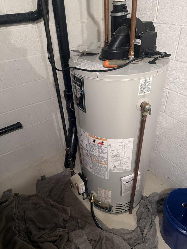 Responded to service call for leaking hot water tank causing property damage. Upon arrival, inspected existing water heater installation and confirmed active leak from aging unit. Documented current setup including power vent configuration with black pump assembly, copper supply lines, and gas connections. Photographed existing Bradford White installation showing vertical black vent piping, copper plumbing connections, and mounting configuration in utility area with concrete block walls. Assessed damage to surrounding area and noted water accumulation at base of unit. Provided detailed estimate for replacement with Bradford White 50-gallon gas tall power vent water heater to match existing specifications and venting requirements.