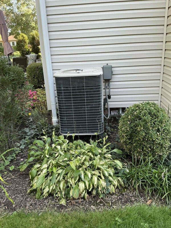 Conducted comprehensive on-site evaluation for complete HVAC system replacement at residential property. Assessed existing outdoor air conditioning condenser unit positioned against exterior wall in landscaped area, documenting current equipment specifications, installation configuration, and surrounding site conditions. Photographed unit placement on concrete pad, electrical disconnect box mounting, refrigerant line routing, and clearance requirements relative to vegetation and structures. Evaluated system accessibility for future replacement installation, noting mulched garden bed with hostas and ornamental plantings requiring protection during equipment changeout. Reviewed electrical service connection and conduit pathways for compatibility with new equipment specifications. Provided detailed replacement options and equipment recommendations based on current system assessment and homeowner requirements.