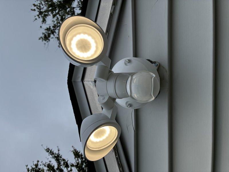 Replaced existing garage floodlight fixture with customer-provided dual-head LED motion sensor security light on exterior metal building. Removed old fixture, installed new mounting hardware, connected electrical wiring to the dual-head unit with central motion detector, and verified proper operation of both LED light heads and motion sensing functionality.