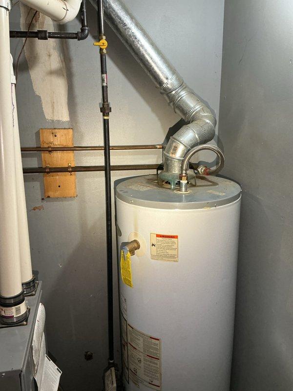 Conducted water heater replacement estimate for residential property. Assessed existing standard white cylindrical tank-type water heater located in basement utility area with visible age-related wear and discoloration. Evaluated current installation including top-mounted exhaust vent with 90-degree metallic ductwork, copper and PVC plumbing connections, gas line with yellow-handled valve, and electrical wiring. Documented equipment specifications, surrounding HVAC infrastructure, and utility room configuration through photographic documentation to provide accurate replacement proposal.
